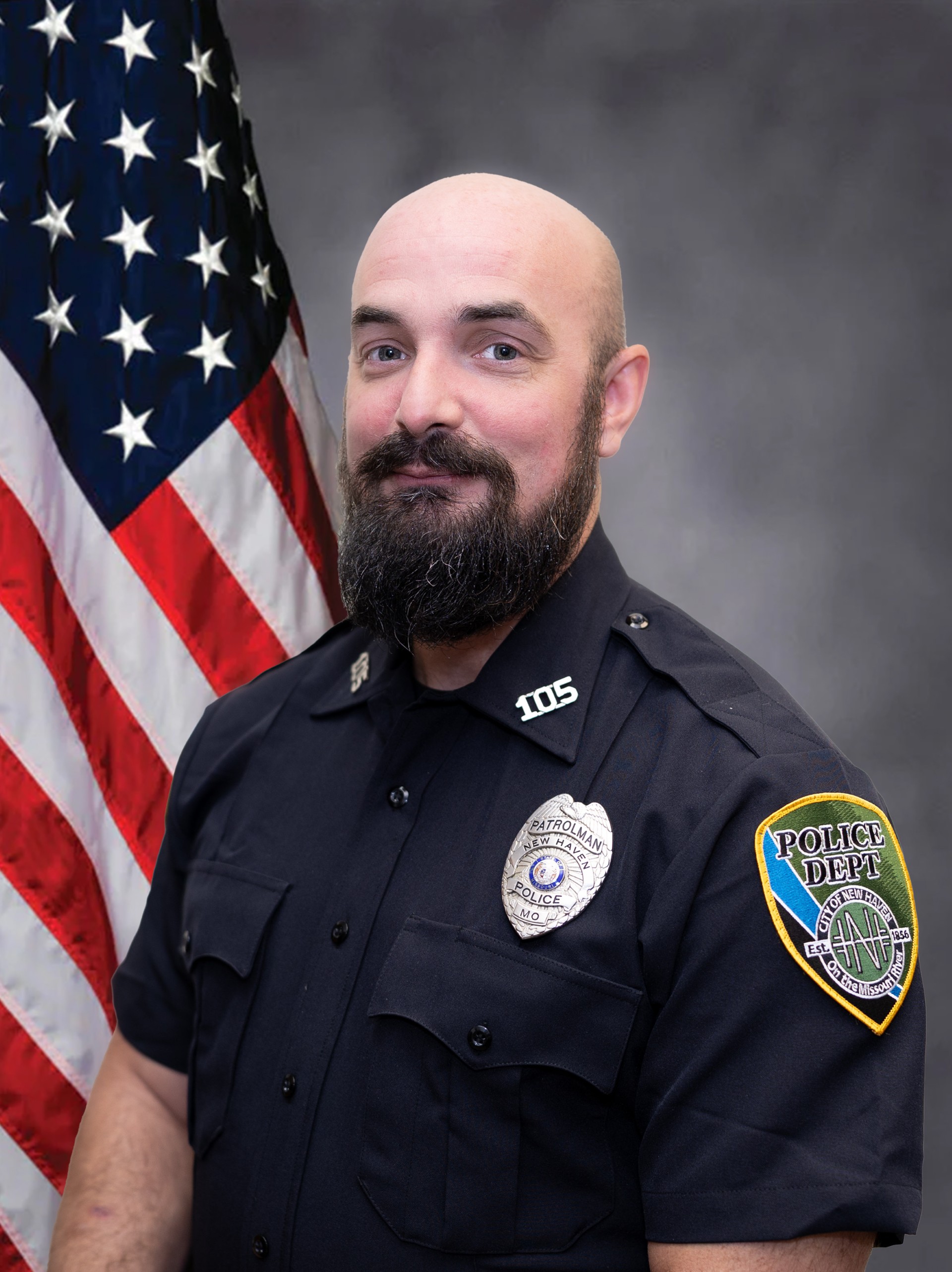 Bald, bearded police officer in a dark uniform with “105” and New Haven Police Dept patches, standing before the U.S. flag.